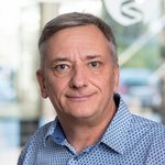 Dr. rer. nat. Peter Heitland steht vor einem Gebäude mit einem grauen und weißen Logo. Er trägt ein kariertes Hemd und hat graue Haare.