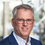 Porträt von Ralf Heitmann, Dipl.-Kfm., in einem Anzug mit offenem Kragen. Hintergrund zeigt unscharfe Büroeinrichtung.