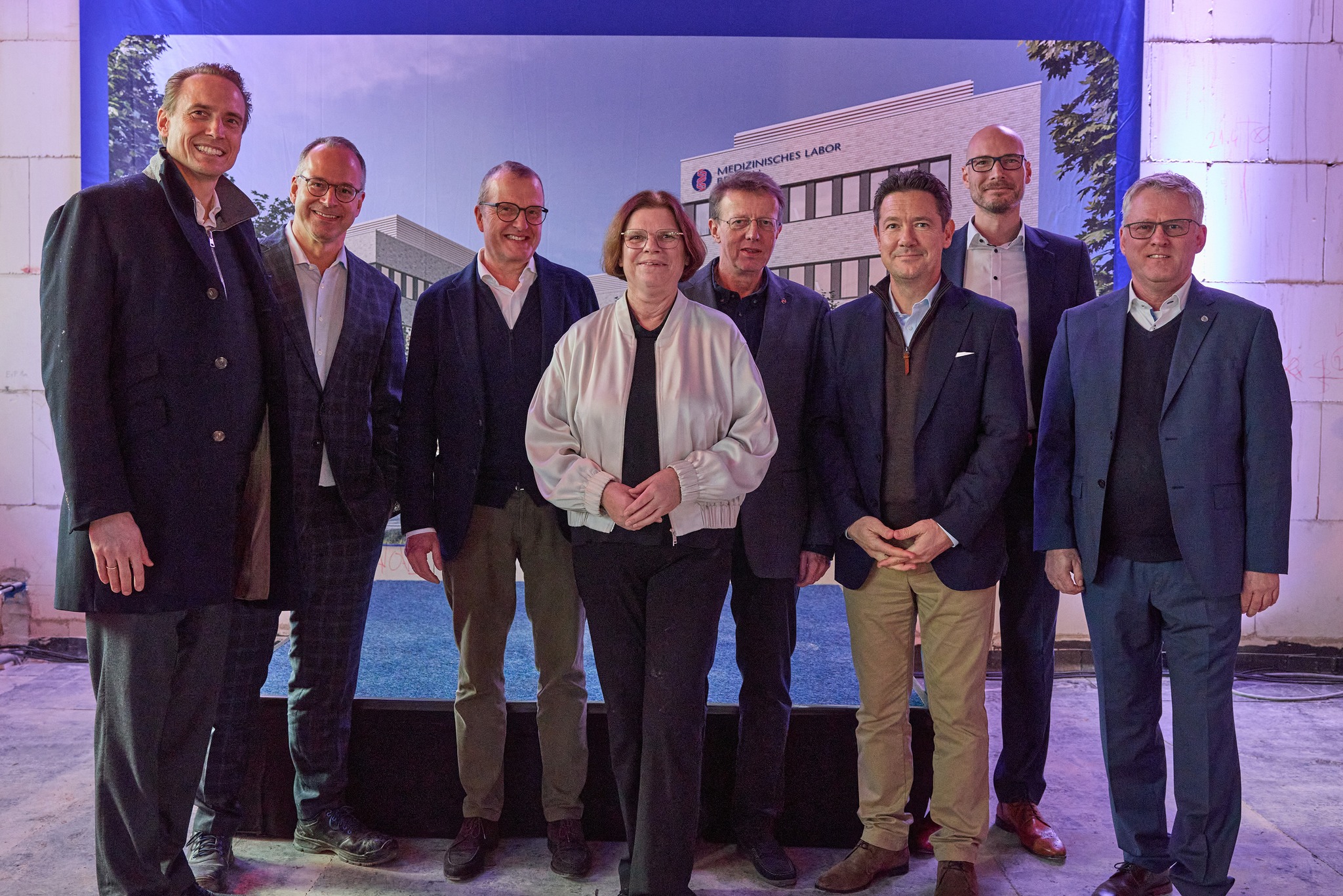 Sieben Personen stehen nebeneinander vor einem Banner mit einem Gebäude und dem Schriftzug 'Medizinisches Labor'. Die Personen tragen formelle Kleidung, darunter Anzüge und eine weiße Jacke.