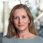 Marina Warnken mit langen, braunen Haaren, trägt ein graues Oberteil. Hintergrund zeigt unscharfe Büroelemente.