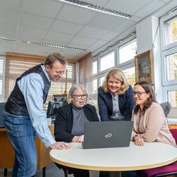Drei Mitarbeiterinnen und ein Mitarbeiter schauen gemeinsam glücklich auf einen Laptop an einem runden Tisch in einem Büro mit großen Fenstern